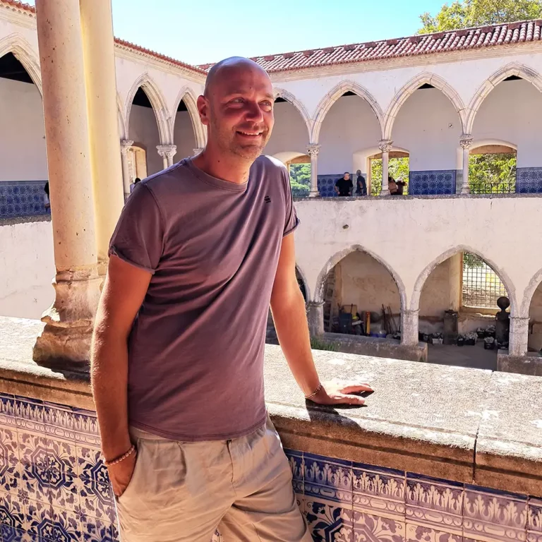 Verslavingscoach Dennis Geelen leunt ontspannen tegen een muurtje in een Mediterraans gebouw.