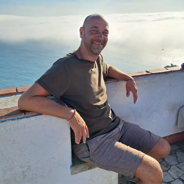 Verslavingscoach Dennis Geelen zit ontspannen tegen een muurtje aan de zee in een Mediterraans landschap.