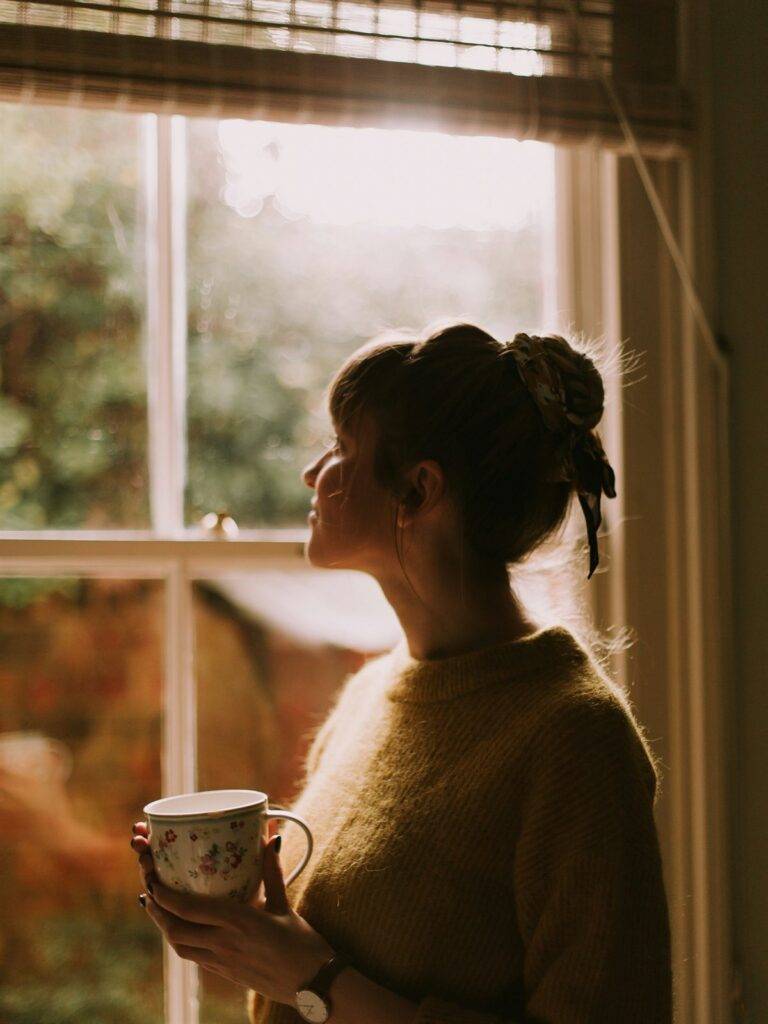 Vrouw staat met een kop koffie bij het raam en kijkt peinzend naar buiten